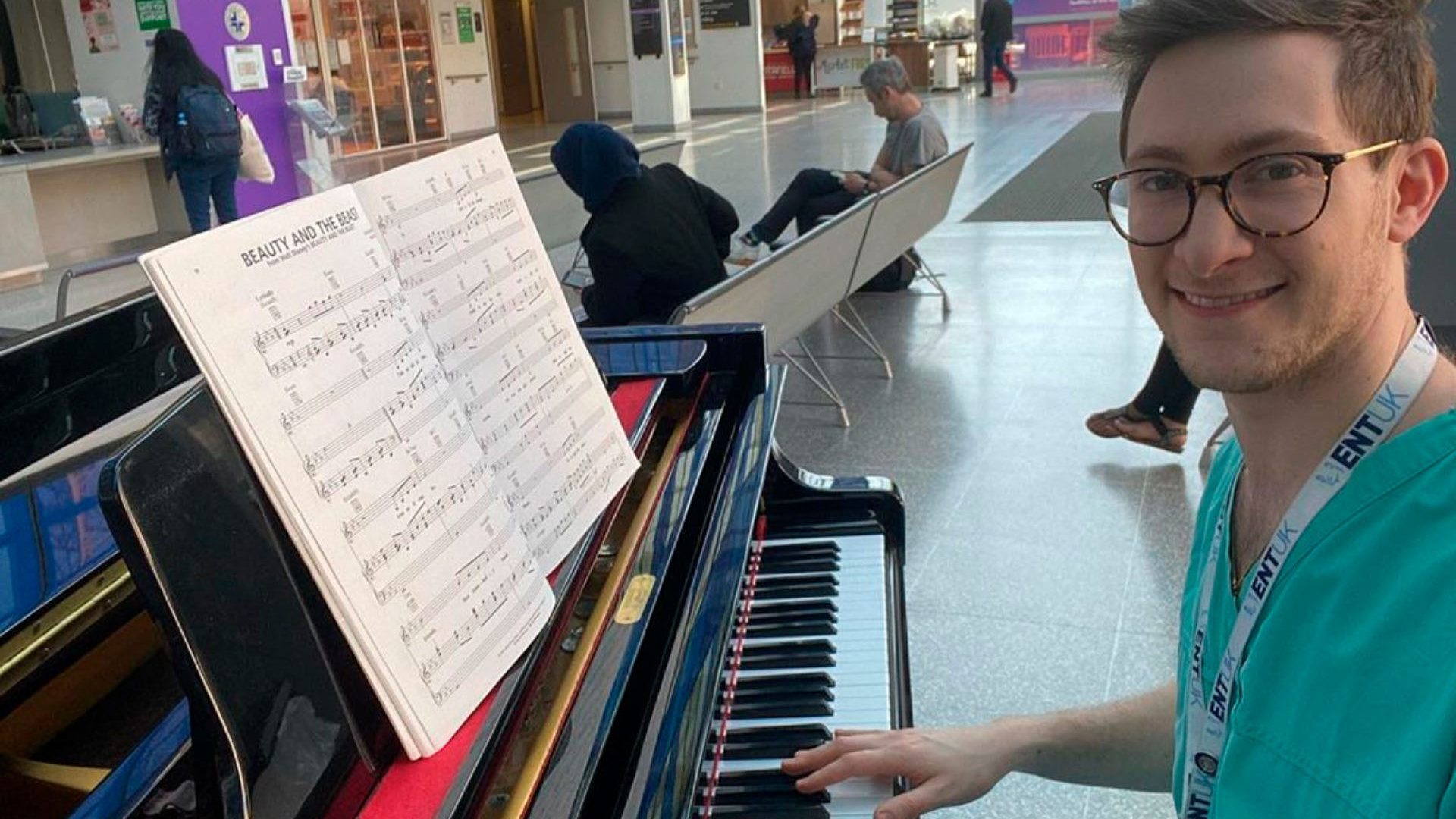 Dr Josh, in team scrubs, sits at a piano with Beauty and the Beast music sheet on the rest