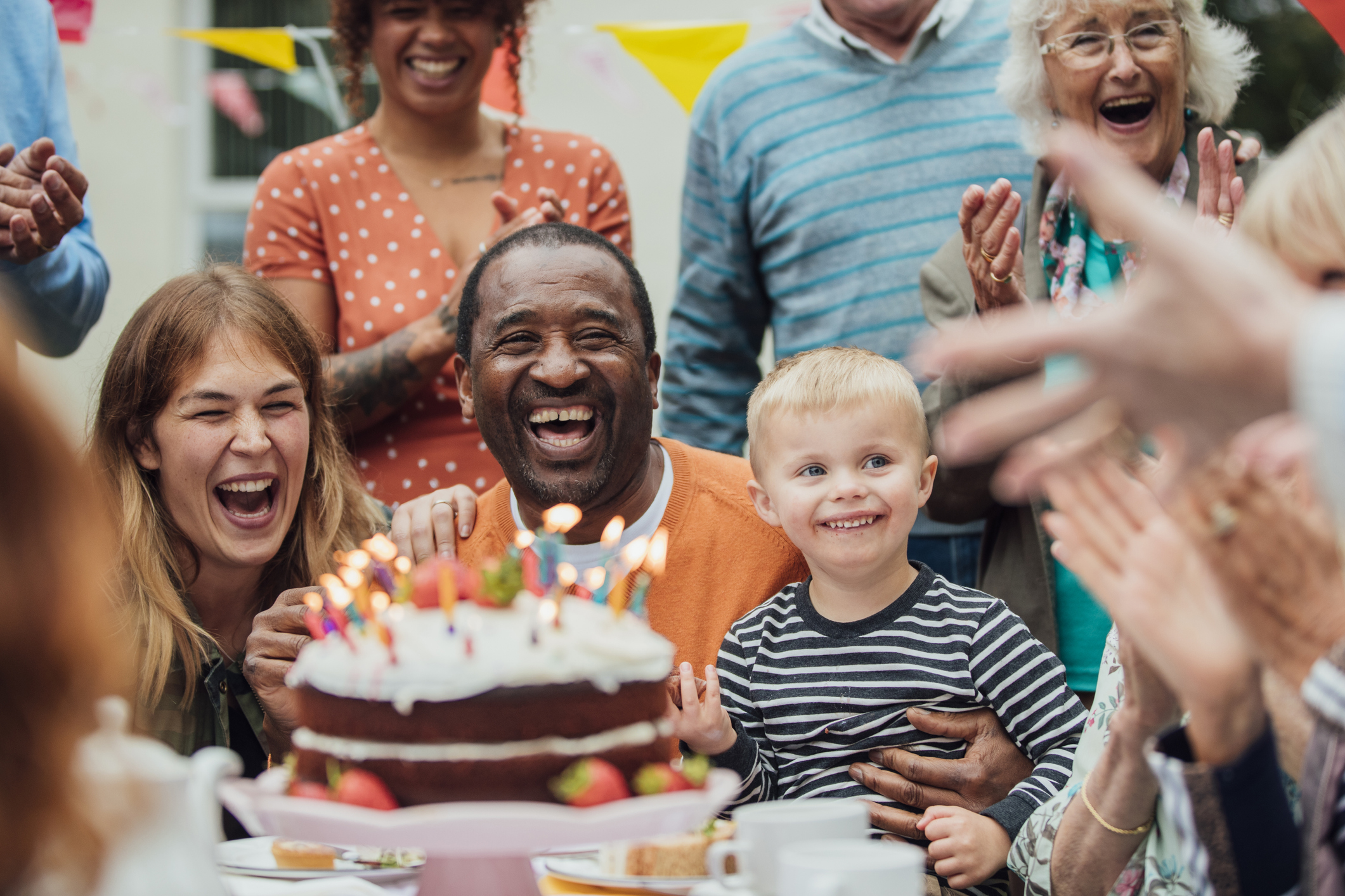 A multi ethnic group of people gather to celebrate a birthday in the garden with lighted candles on a cake. Everyone is laughing and clapping together.