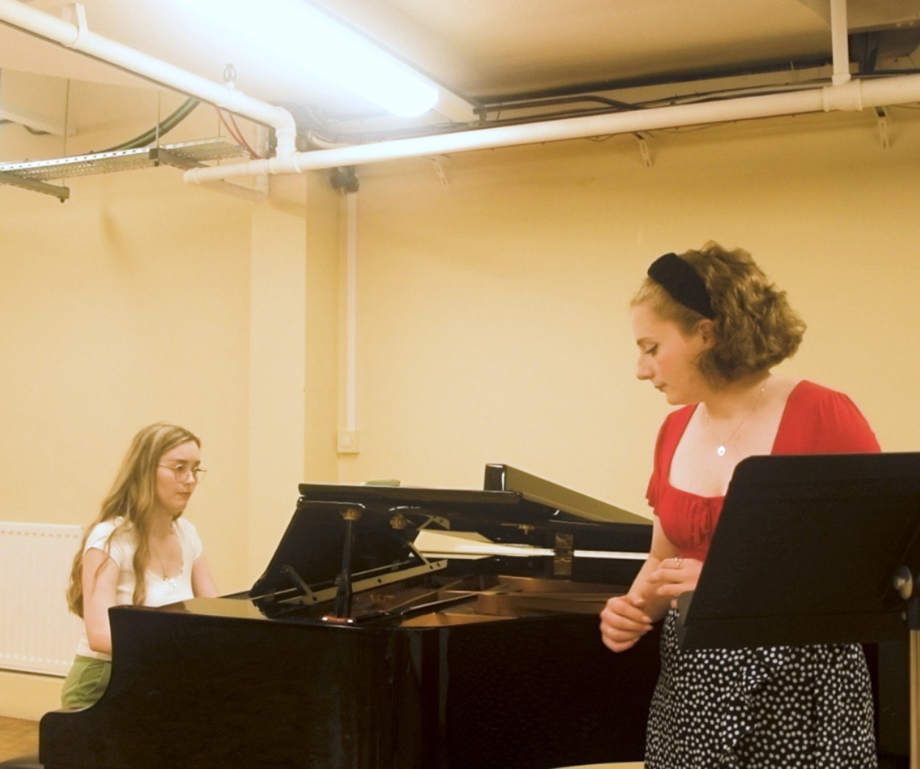 Two women in a room, woman on the left with blond hair and white t-shirt is playing the piano sat down, while the other one is on the right side, leaning on the piano, with short brown hair and a red t-shirt