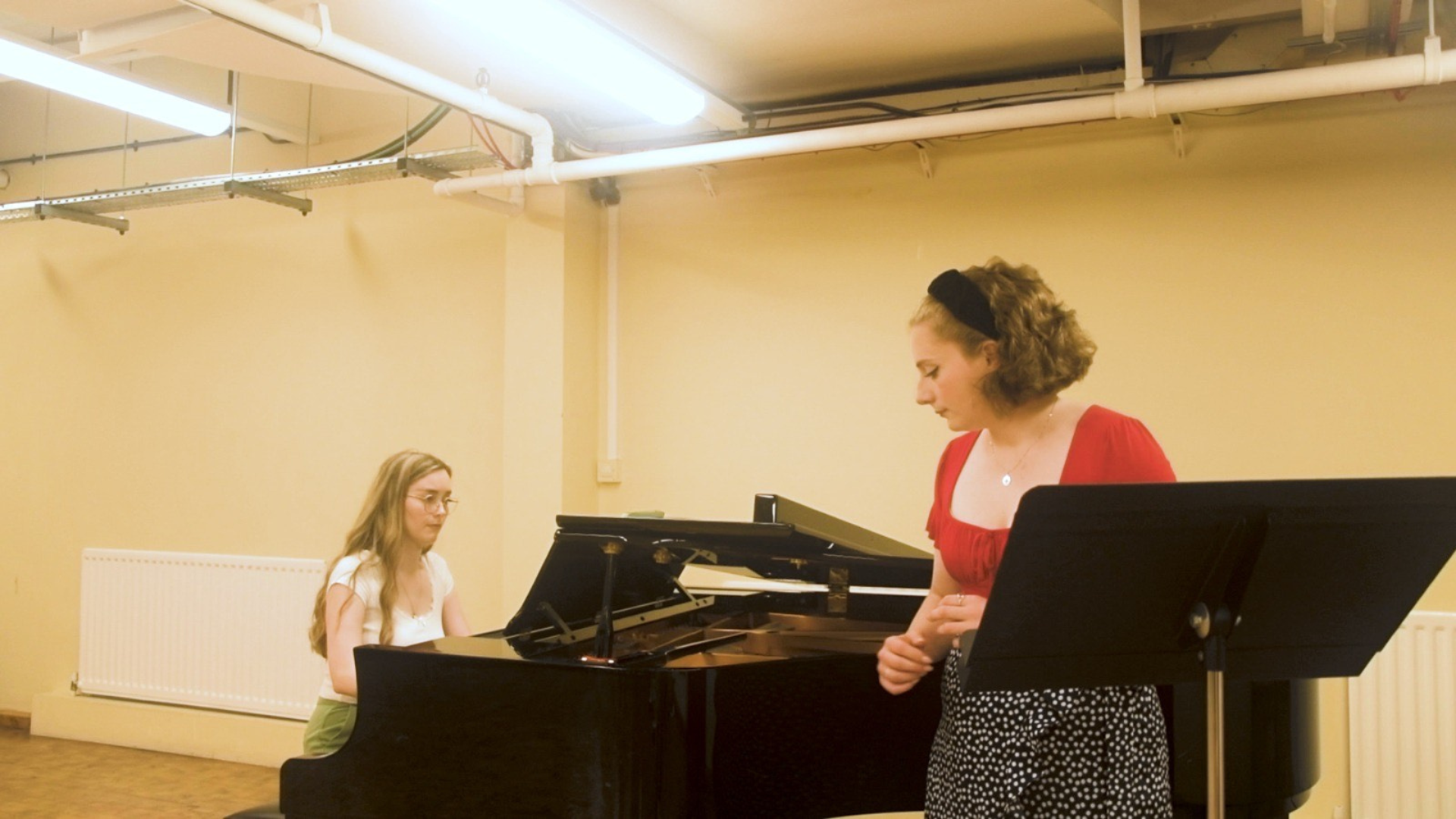 Two women in a room, woman on the left with blond hair and white t-shirt is playing the piano sat down, while the other one is on the right side, leaning on the piano, with short brown hair and a red t-shirt