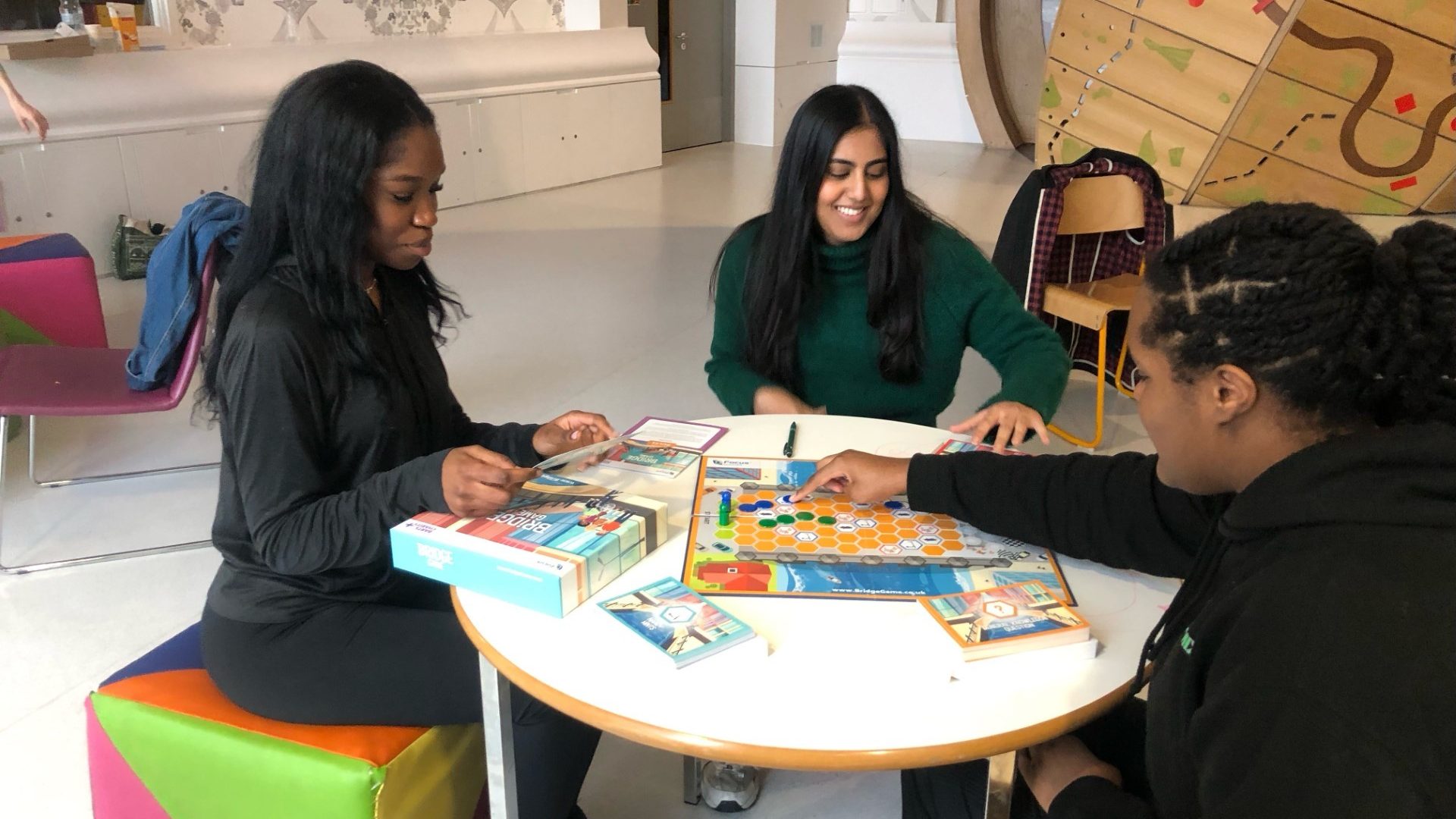 Three young adults sit around a white table playing the board game called Bridge Game