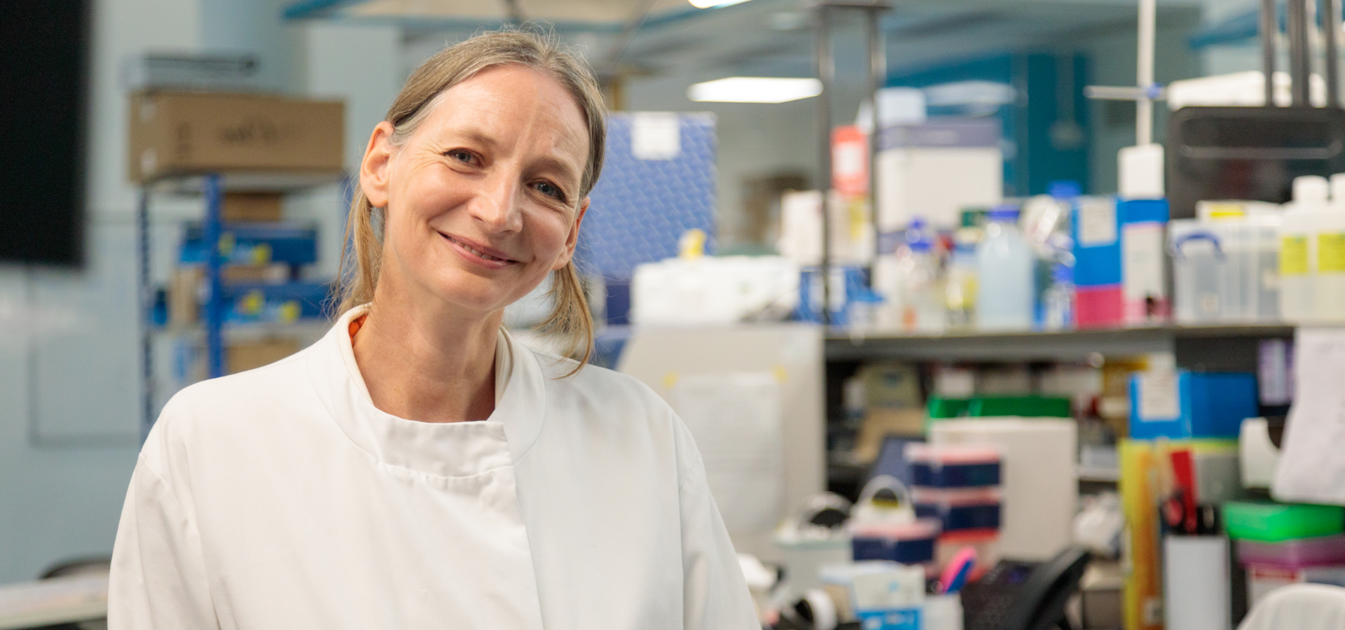Researcher Cleo Bishop photographed in a lab coat, smiling to camera in a lab setting.