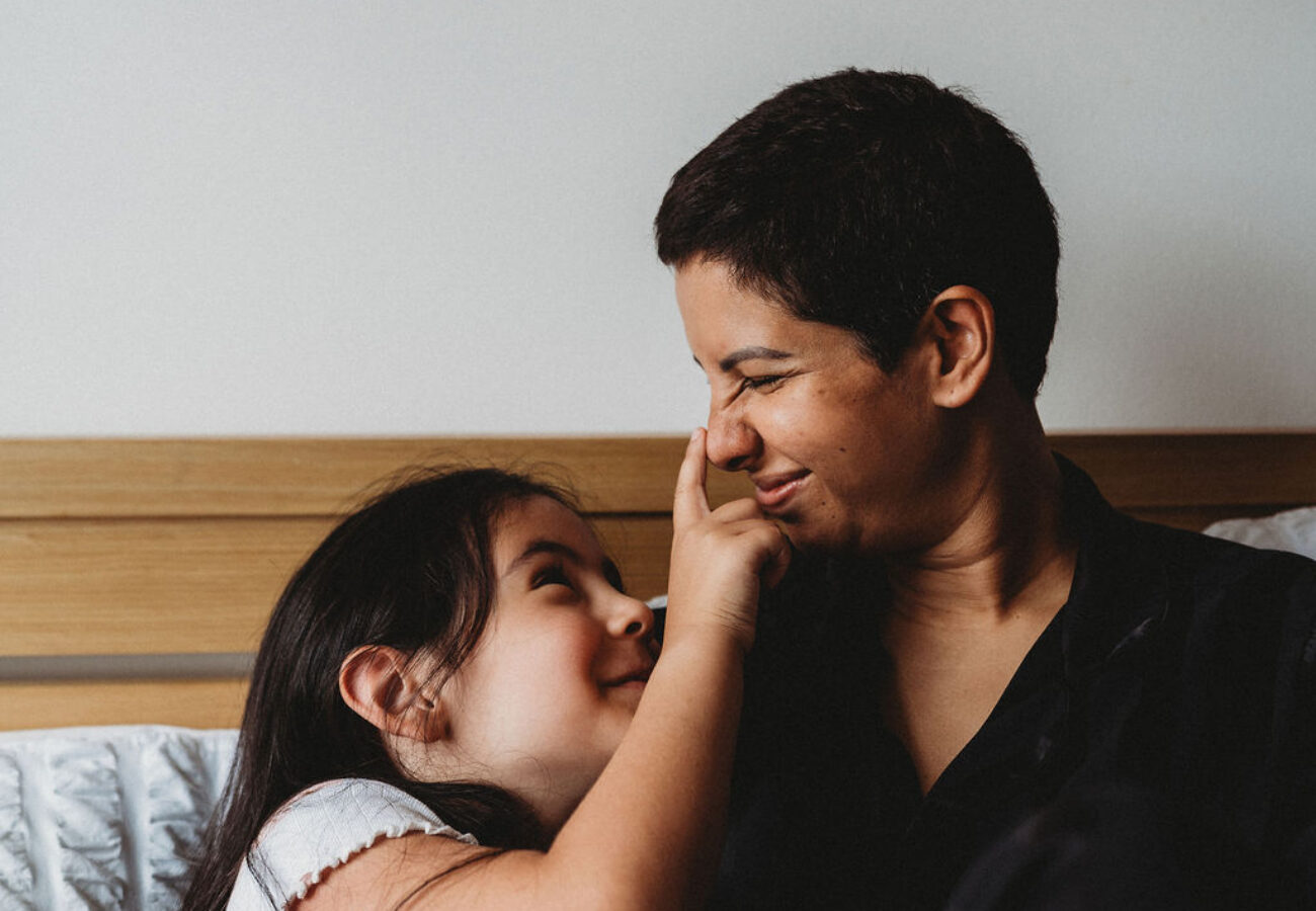 A woman and her daughter sat on a bed, the daughter has playfully pressed her mum's nose, both are smiling