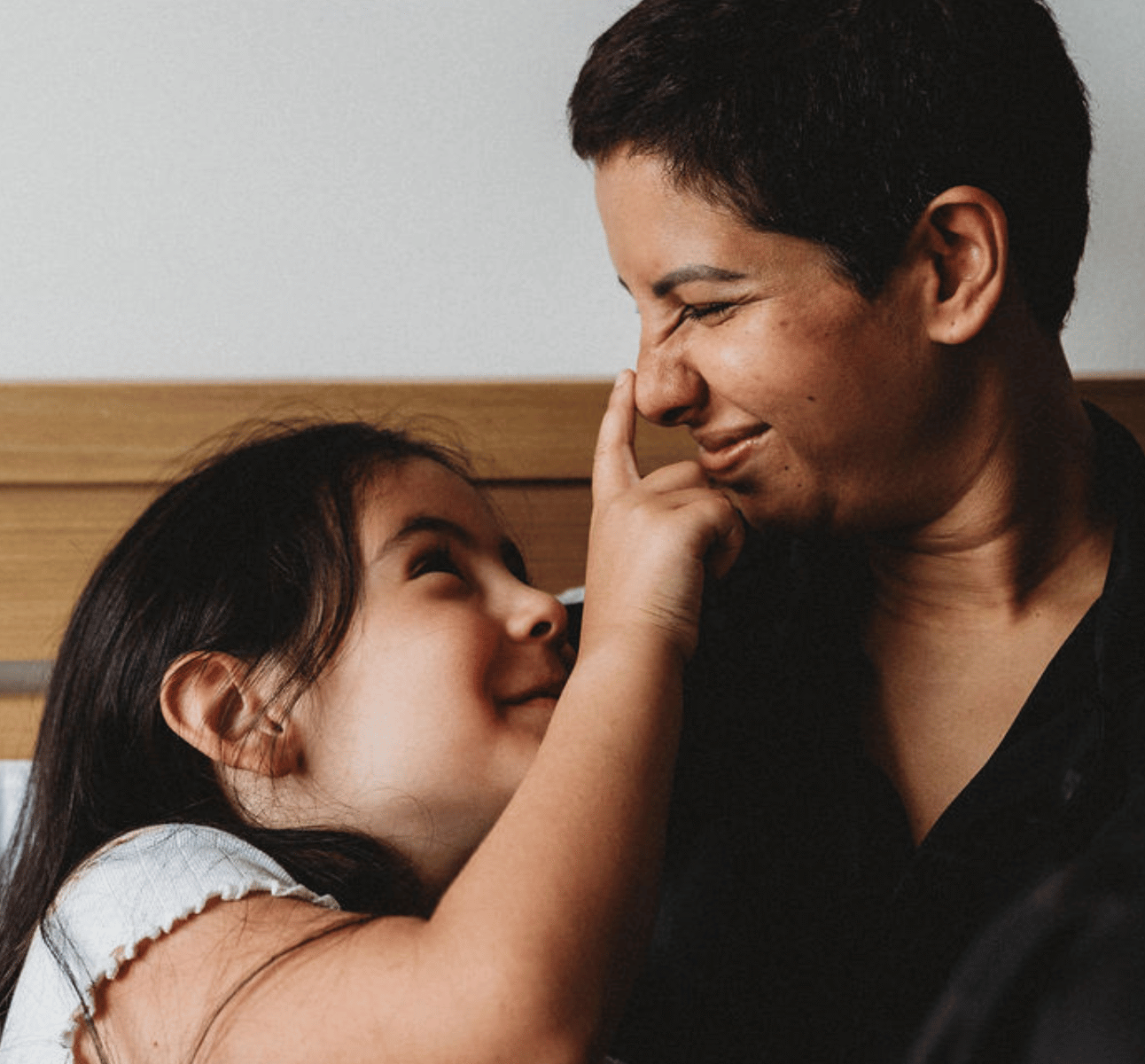 A woman and her daughter sat on a bed, the daughter has playfully pressed her mum's nose, both are smiling