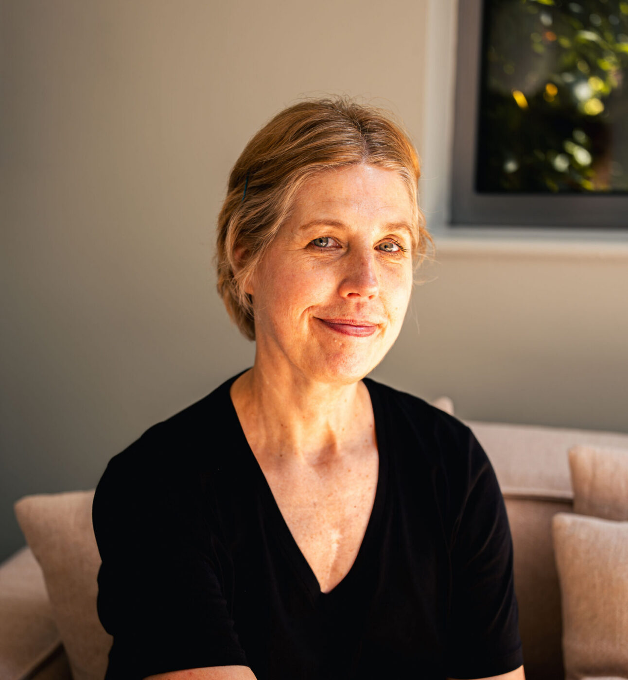 A woman in a black t-shirt smiling at the camera, sat on a couch.