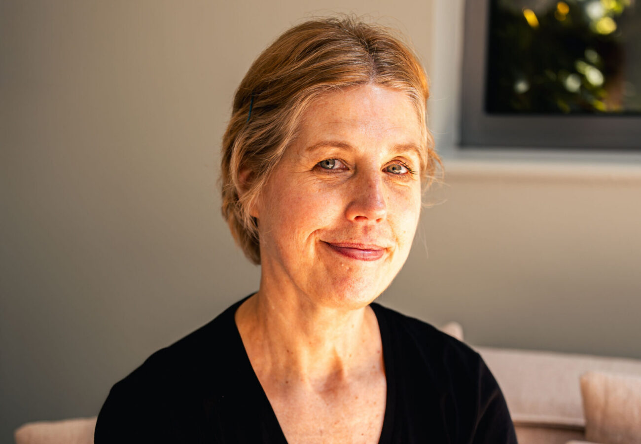 A woman in a black t-shirt smiling at the camera, sat on a couch.