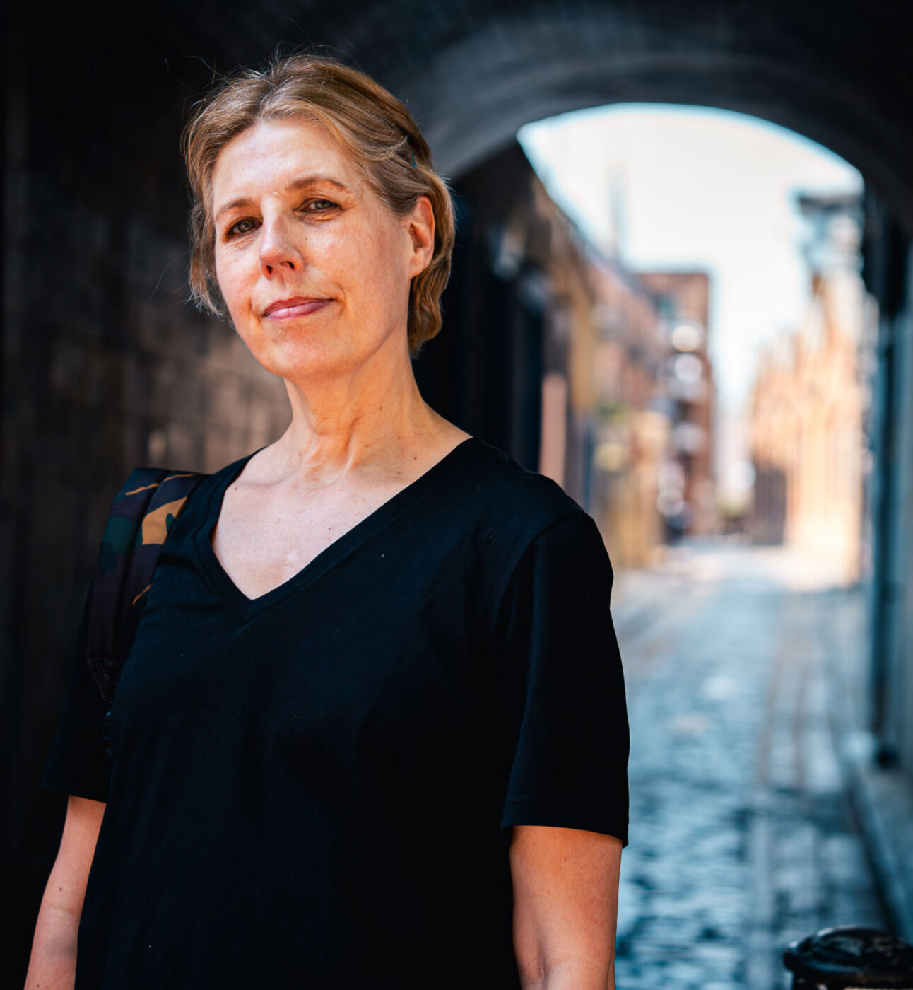 A woman standing in an alley, smiling to the camera wearing a black tshirt