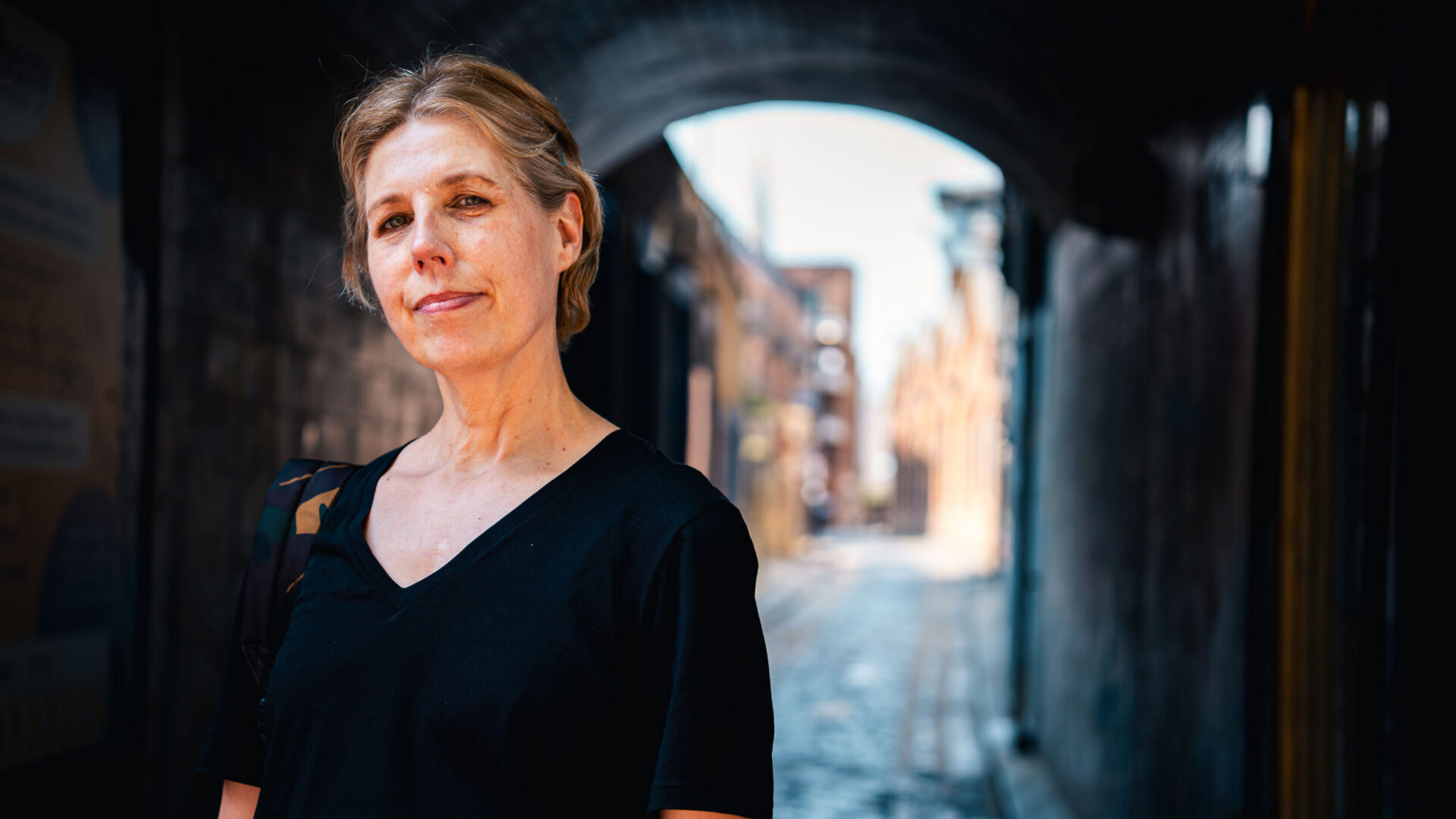 A woman standing in an alley, smiling to the camera wearing a black tshirt