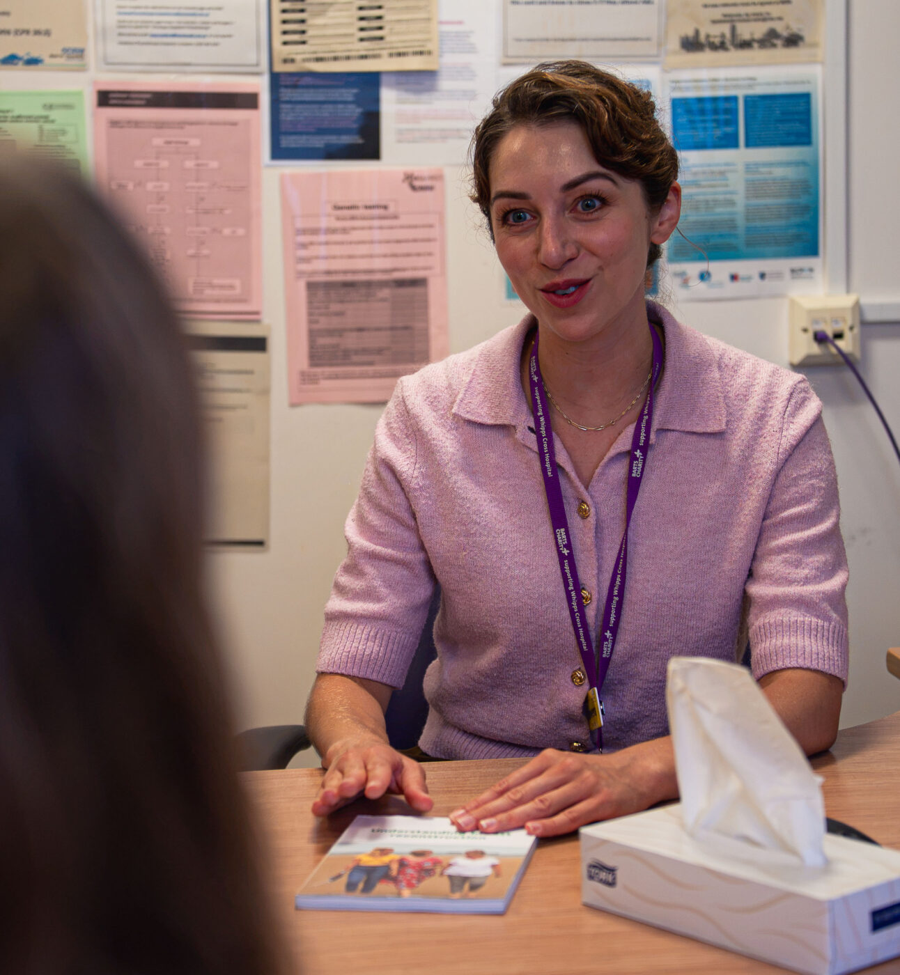A woman in a pink shirt with a lanyard sitting behind a desk in what seems to be an office, there is tissue in front of her and someone on the other side she's talking to. She seems to be talking to them.