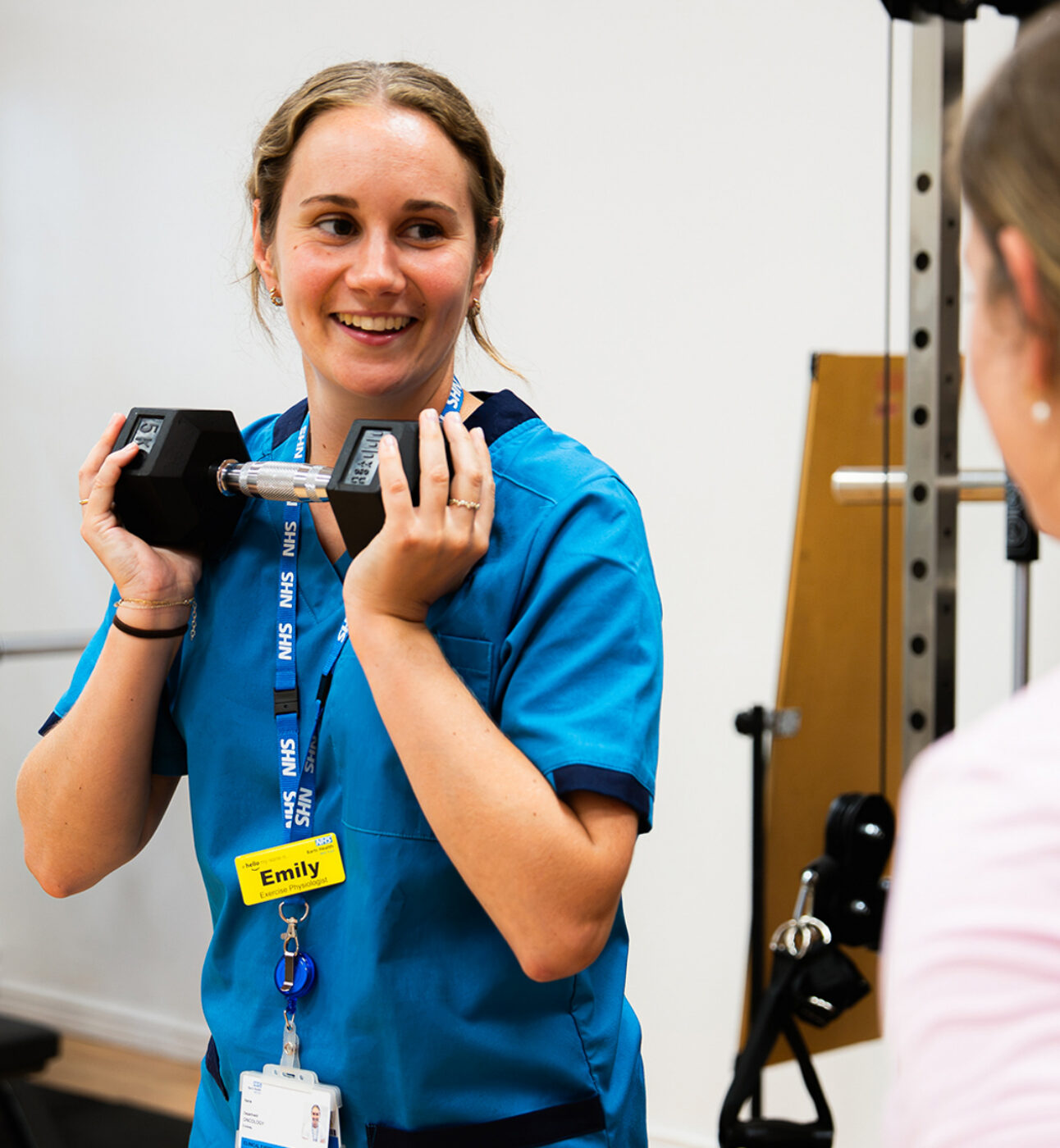 A woman in a medical uniform standing in front of another woman showing her how to do an exercise with a dumbbell. The nurse is holding the dumbbell to her chest and smiling
