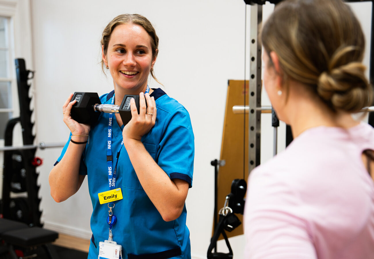 A woman in a medical uniform standing in front of another woman showing her how to do an exercise with a dumbbell. The nurse is holding the dumbbell to her chest and smiling