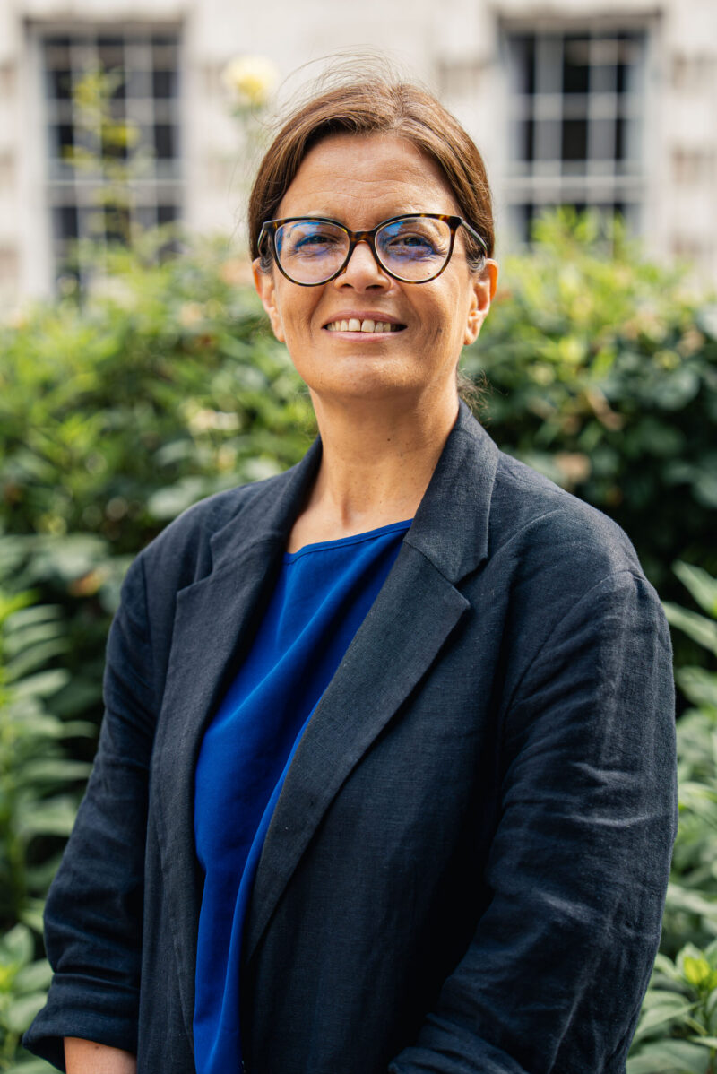 A woman wearing a black blazer wearing glasses looking into the camera, standing in front of some greenery.