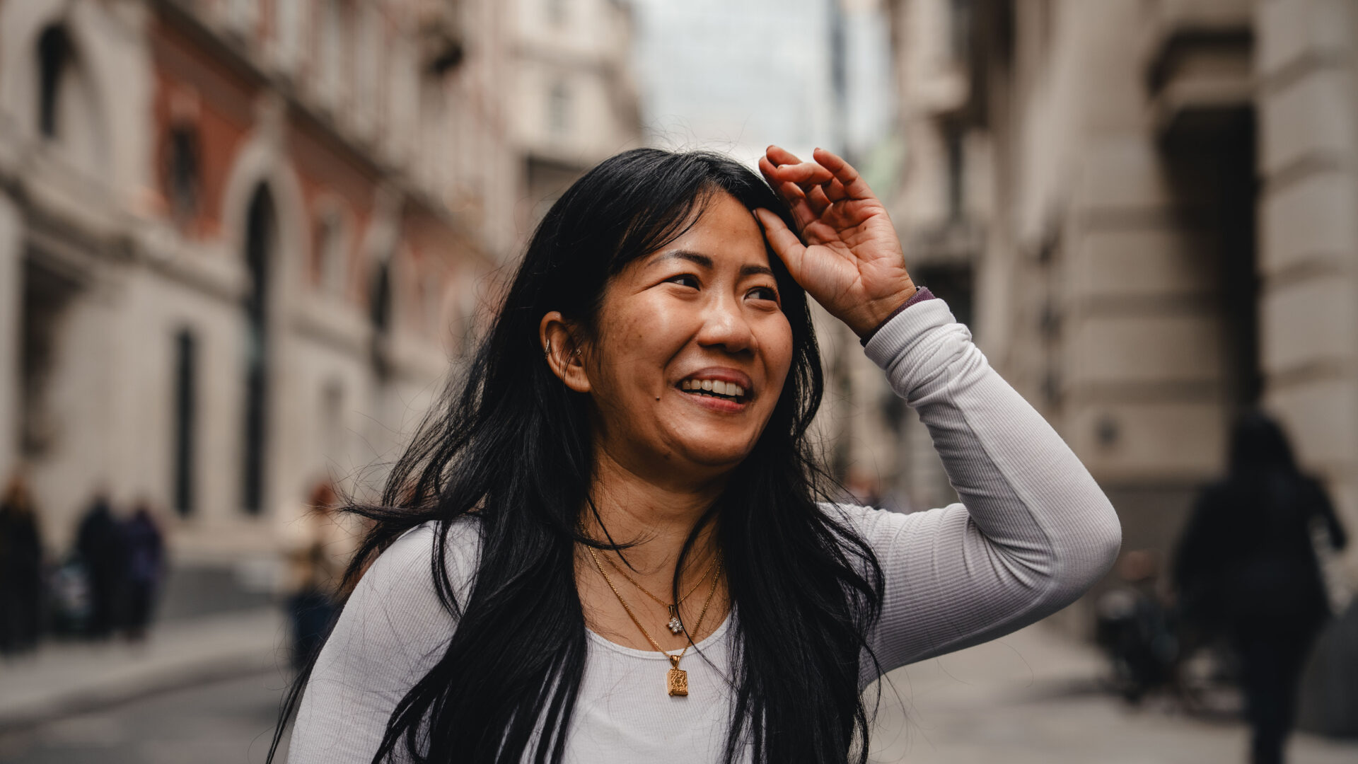 A woman photographed in an urban setting, she's smiling looking away from the camera, one hand is pushing hair away from her face gently