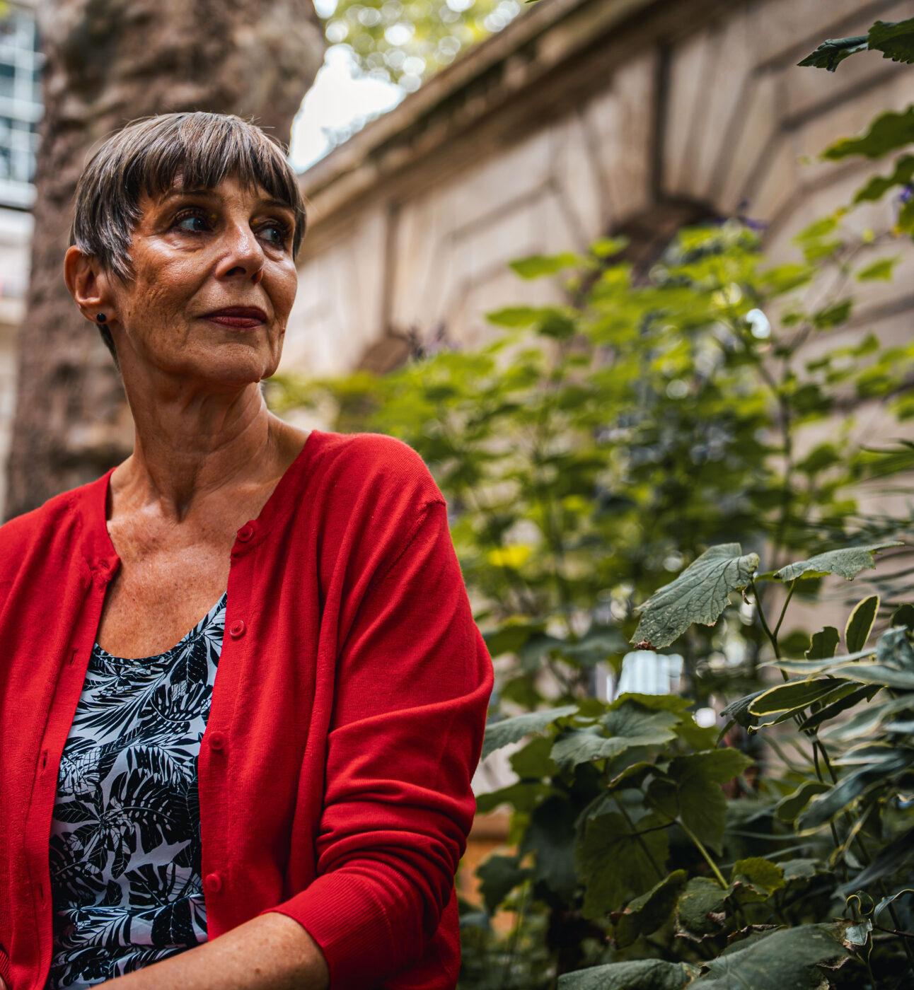 A woman in a bright red cardigan and short hair sitting in what looks like to be a park or a garden, looking into the distance.