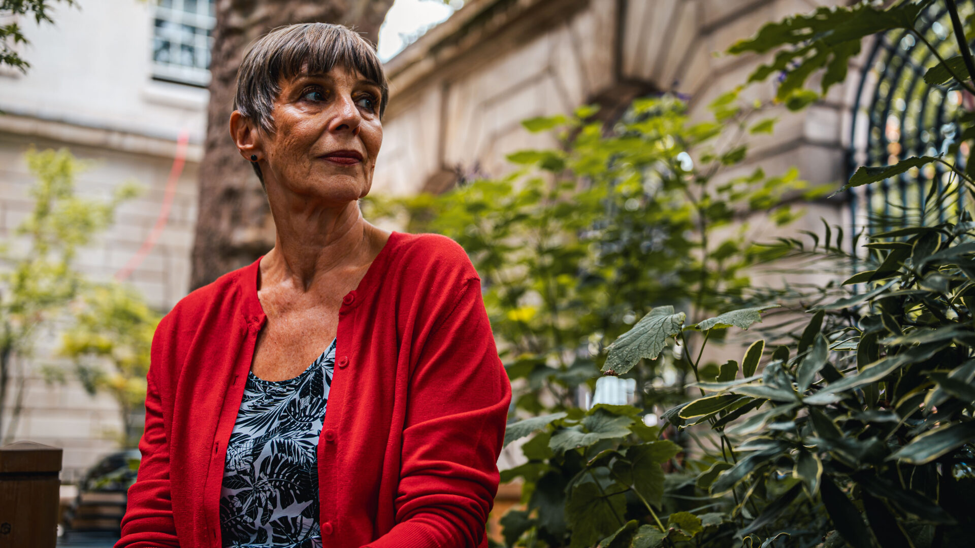 A woman in a bright red cardigan and short hair sitting in what looks like to be a park or a garden, looking into the distance.