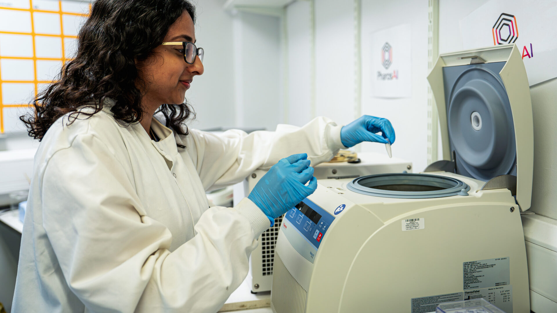 A female researcher in a lab, wearing a lab coat and lab gloves, holding a pipette. Research lab in the background.