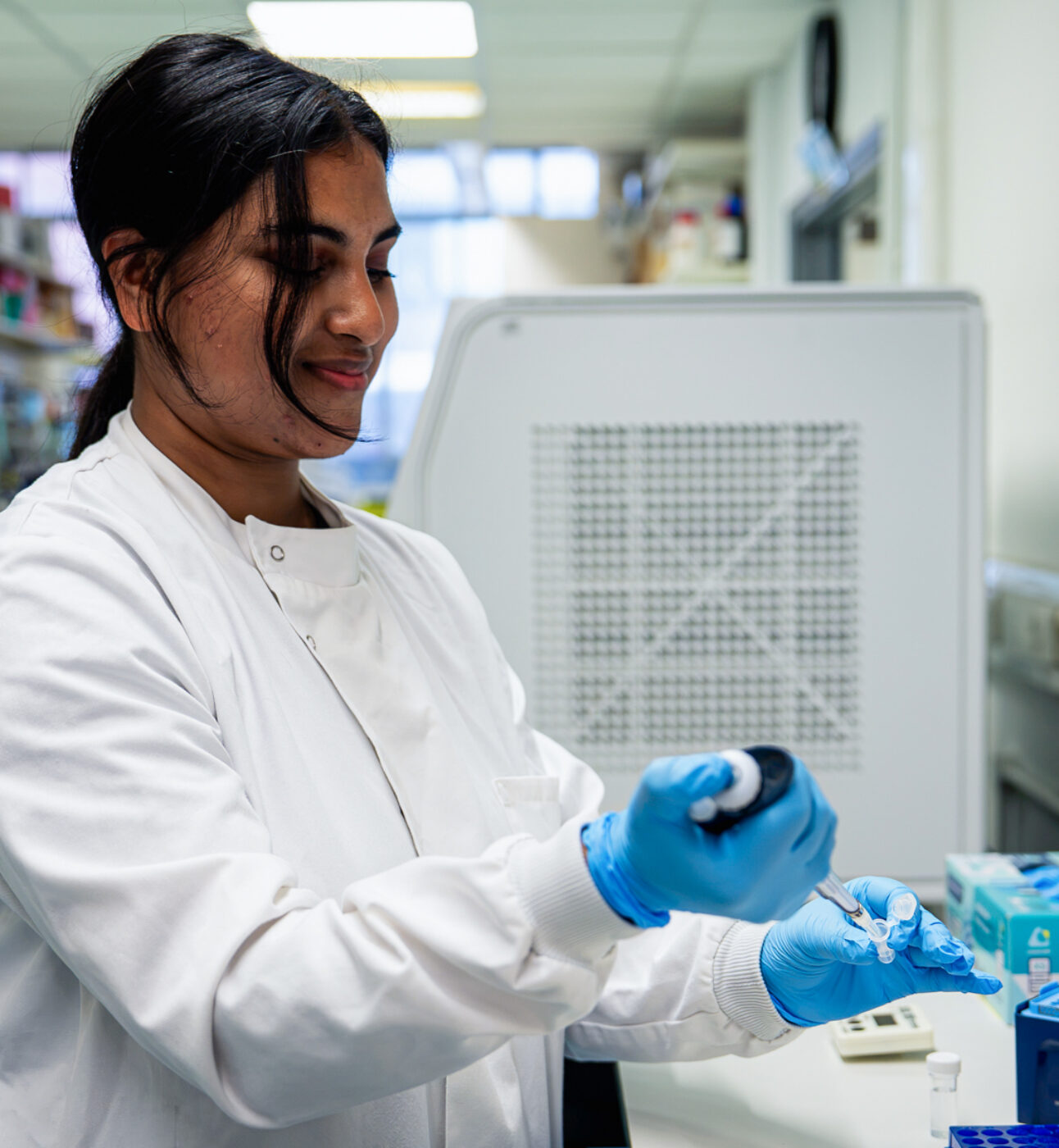 A female researcher in a lab, wearing a lab coat and lab gloves, holding a pipette. Research lab in the background.