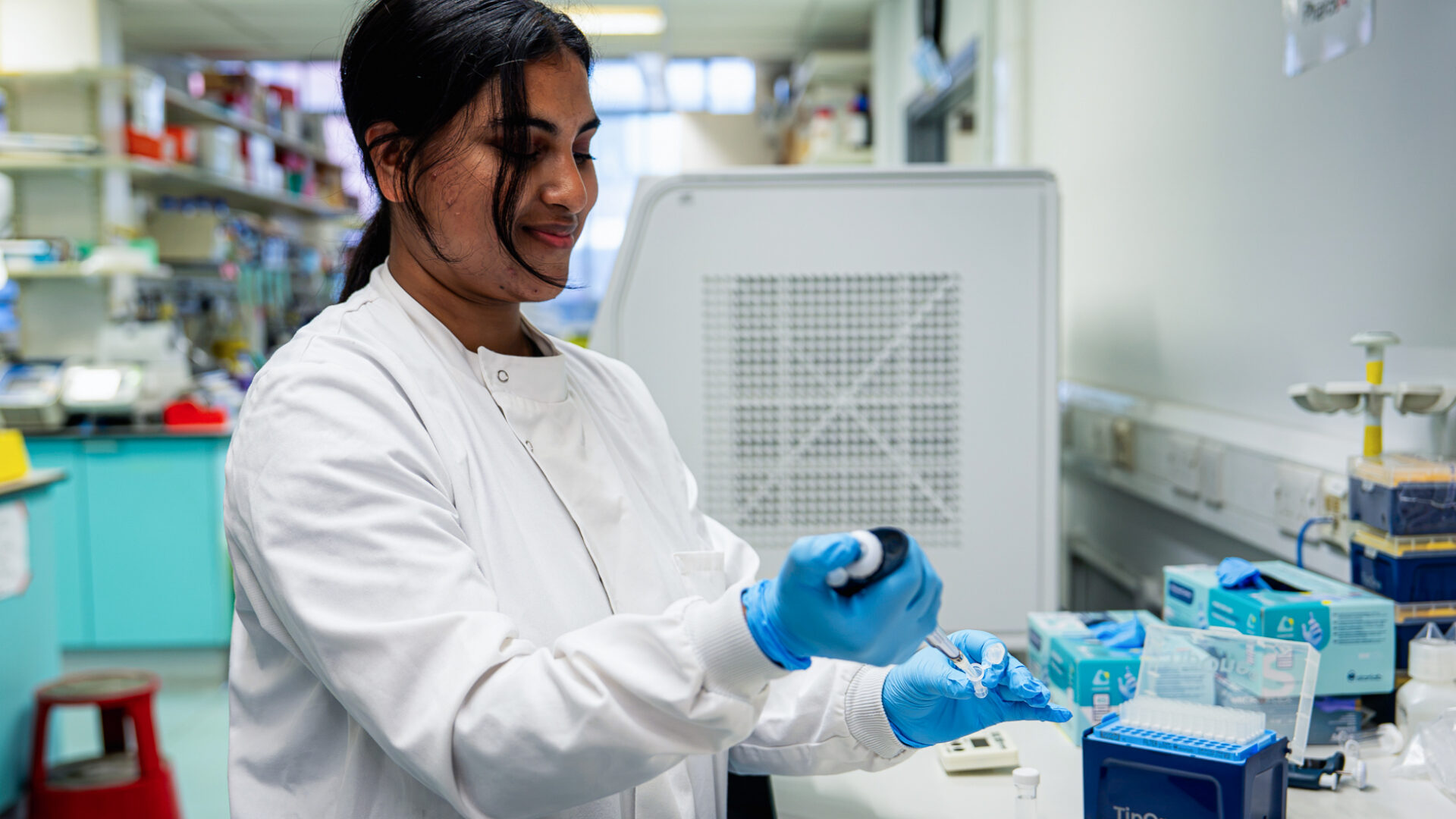 A female researcher in a lab, wearing a lab coat and lab gloves, holding a pipette. Research lab in the background.