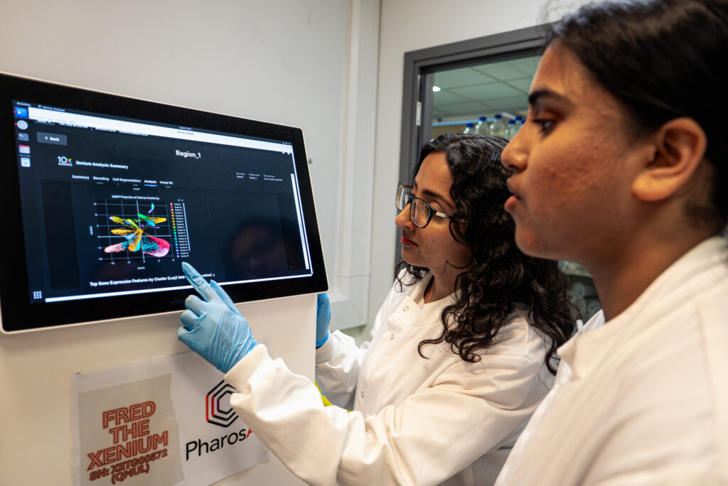 Two female researchers looking at what seems to be research information on a screen, one of them is pointing at it, her hand is in a surgical glove. THey seem to be having a conversation about the screen.