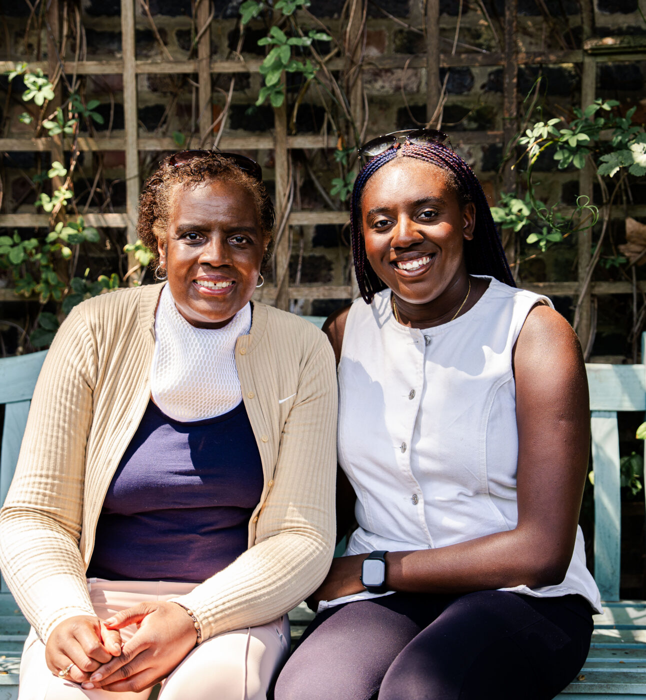 Two women sitting on a park bench very close to each other, they look related perhaps mother and daugher, both looking at the camera smiling
