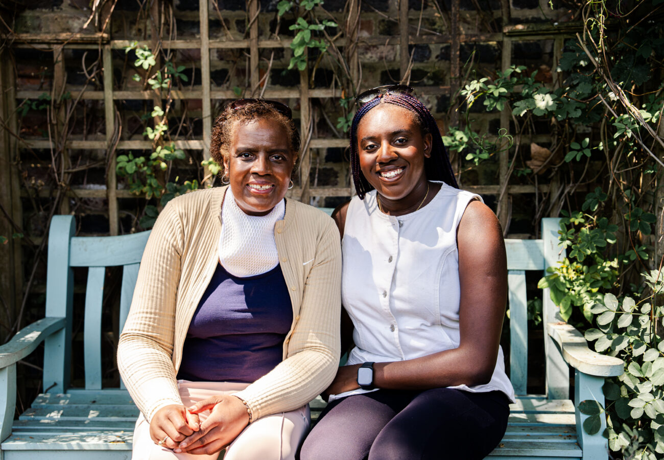 Two women sitting on a park bench very close to each other, they look related perhaps mother and daugher, both looking at the camera smiling