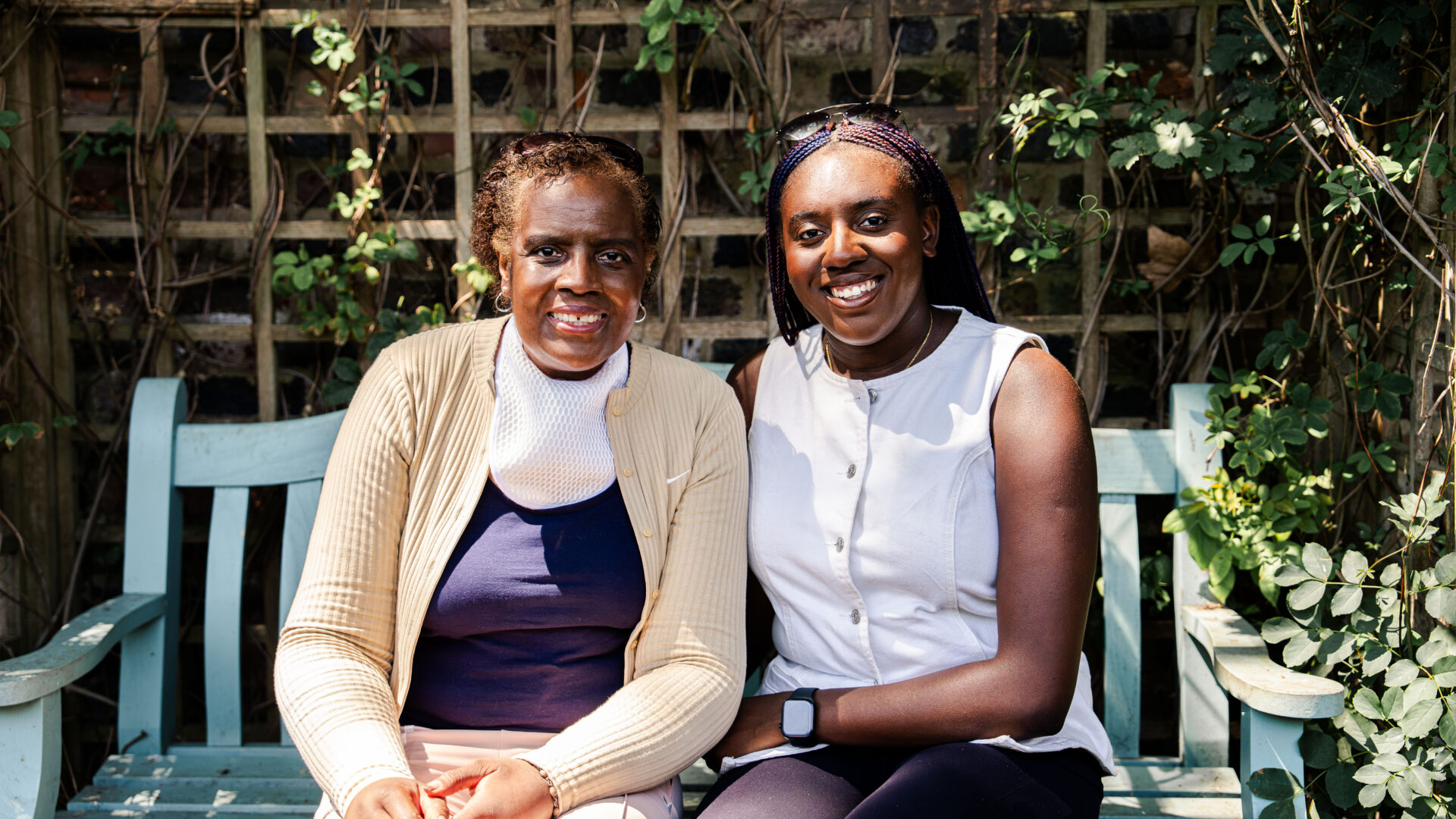 Two women sitting on a park bench very close to each other, they look related perhaps mother and daugher, both looking at the camera smiling