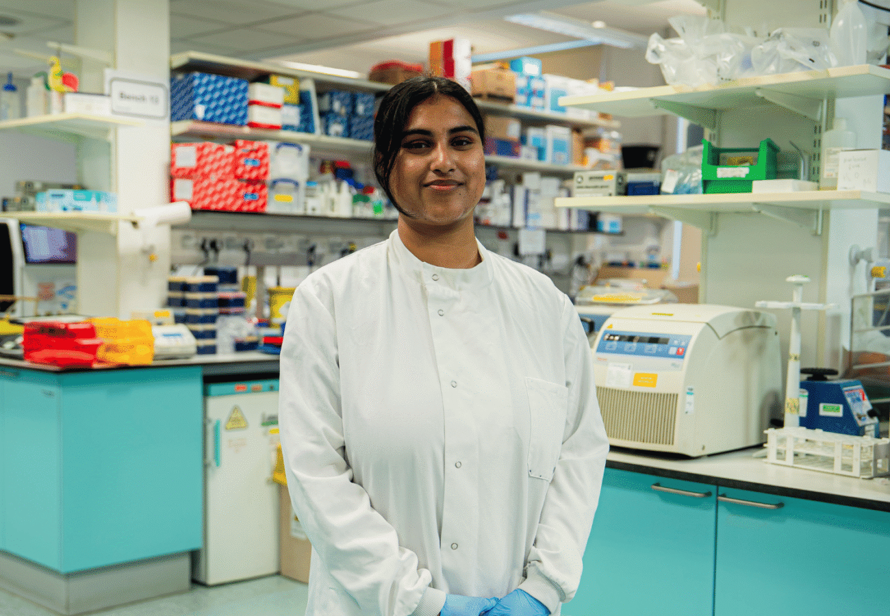 A woman standing in a lab looking at the camera, she's wearing a lab coat and surgical gloves with her hands in front of her body.
