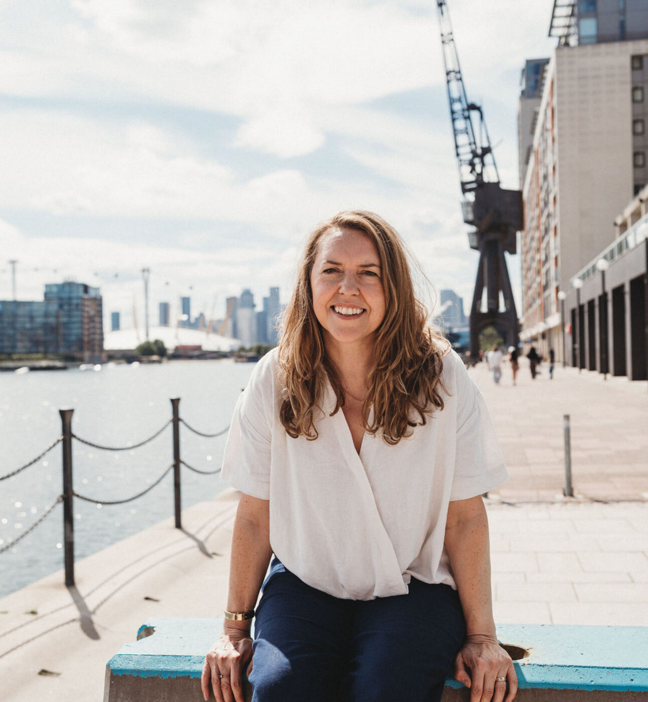 A woman with long blond hair leaning on a wall at what seems to be a dock or a port, there's water behind her and a city skyline. She's looking into the camera smiling.